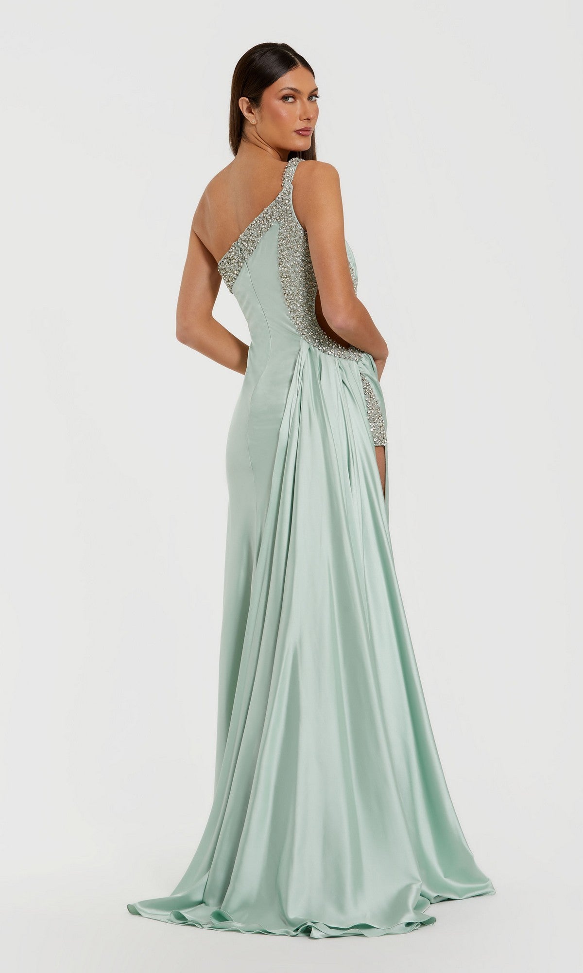 A woman stands and looks over her shoulder in the Mac Duggal 68447 long formal dress—a light green, one-shoulder satin gown featuring sequin accents and a draped train.